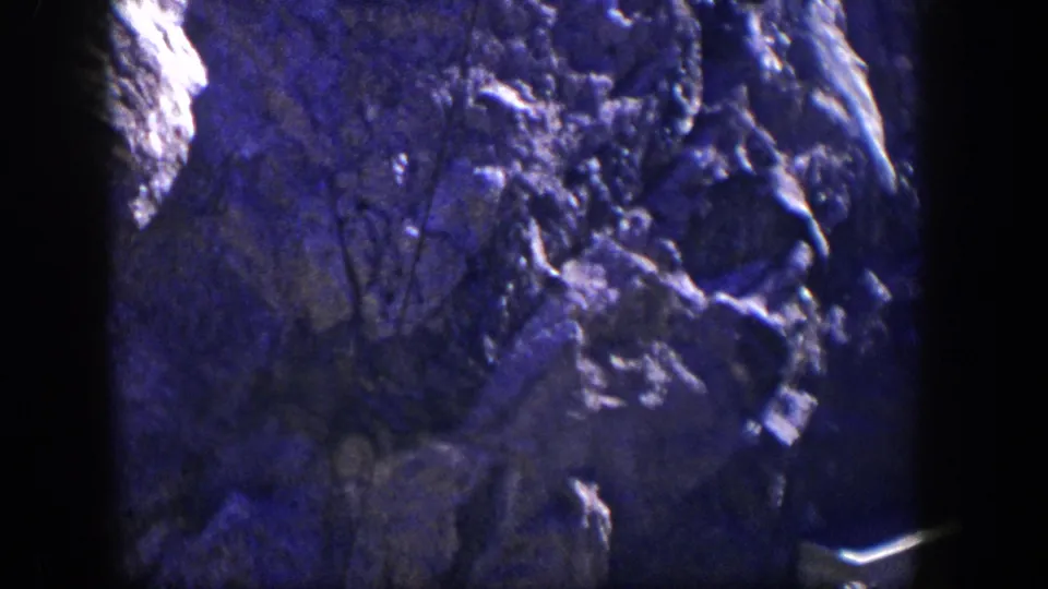 1954 Nevada 8mm frame with a rocky cliff surface, brush silhouettes, dark film border, and a small pale structure near the lower right.