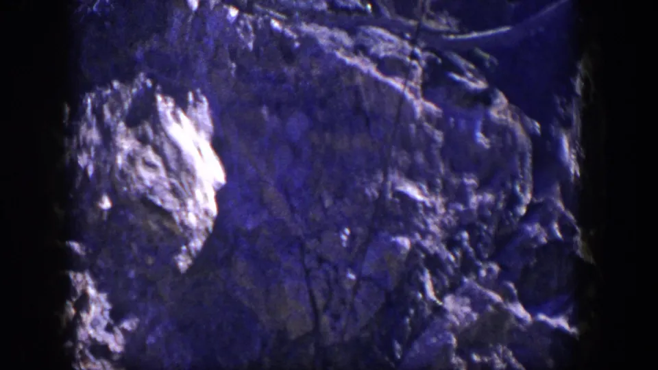 1954 Nevada 8mm frame of a rugged cliff face with bright rock patches, dark cracks, sparse vegetation, and visible film-edge borders.