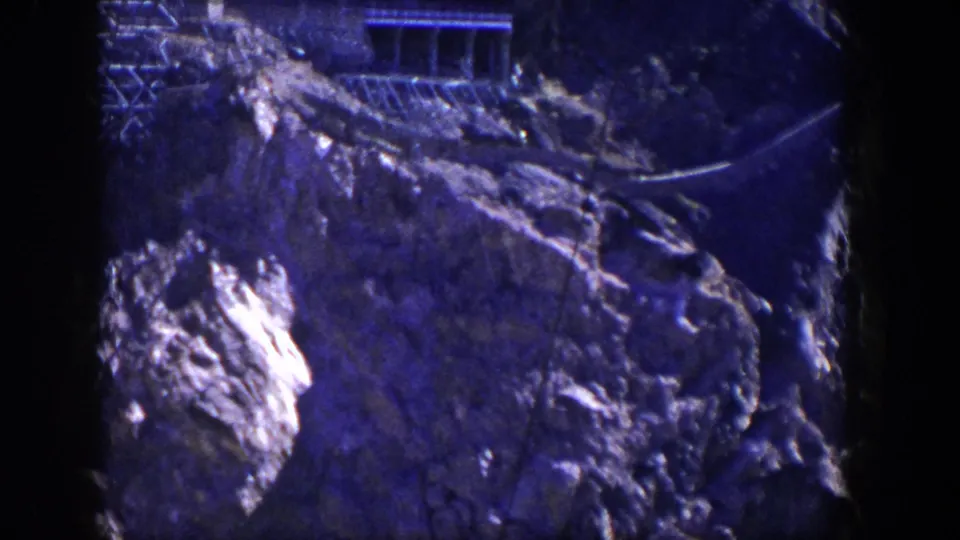 1954 Nevada 8mm frame showing a sunlit rocky slope with a building or platform near the upper edge.
