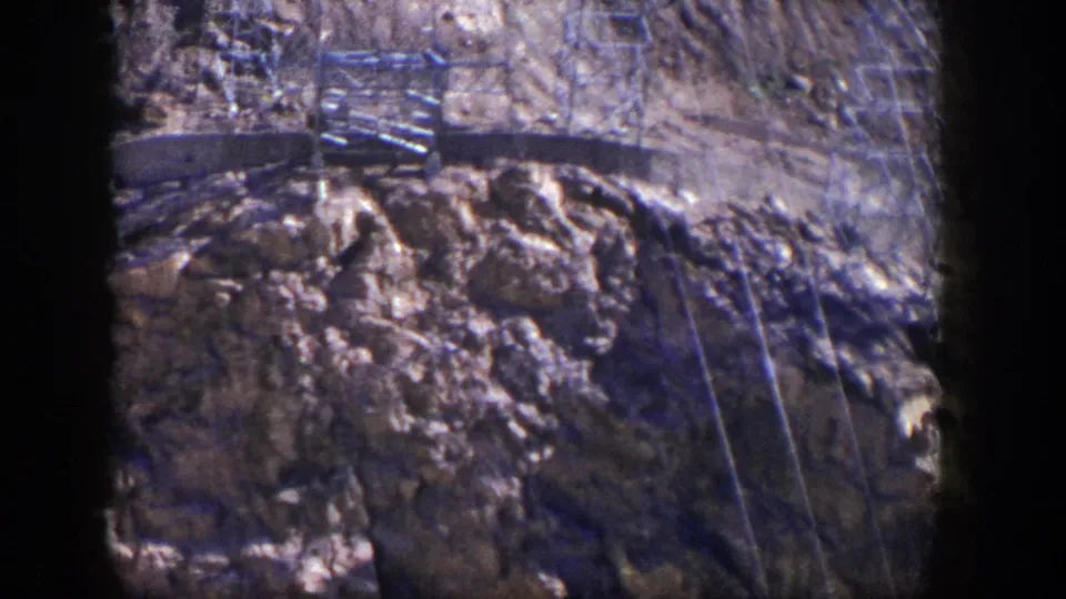 Transmission tower forms and power lines stand above rugged tan rocks in a 1954 Nevada home-movie view.
