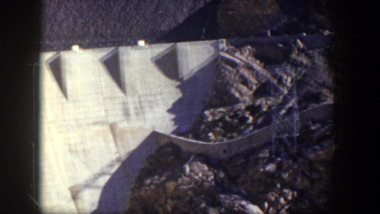 1954 Nevada 8mm frame showing a large concrete dam face, curved edge, dark rock, and strong shadow across the structure.
