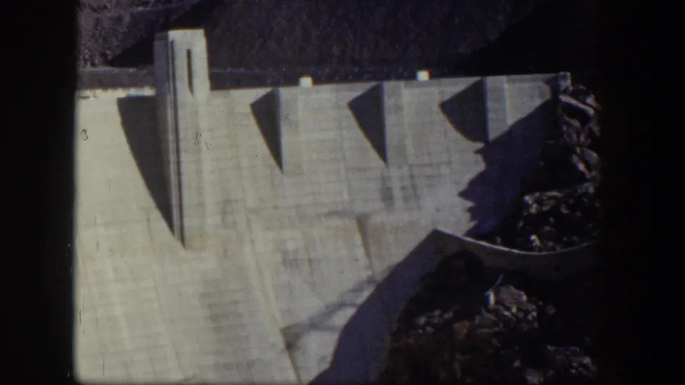 1954 Nevada 8mm frame of the dam wall, a tall vertical structure, angular shadows, and rocky terrain along the side.