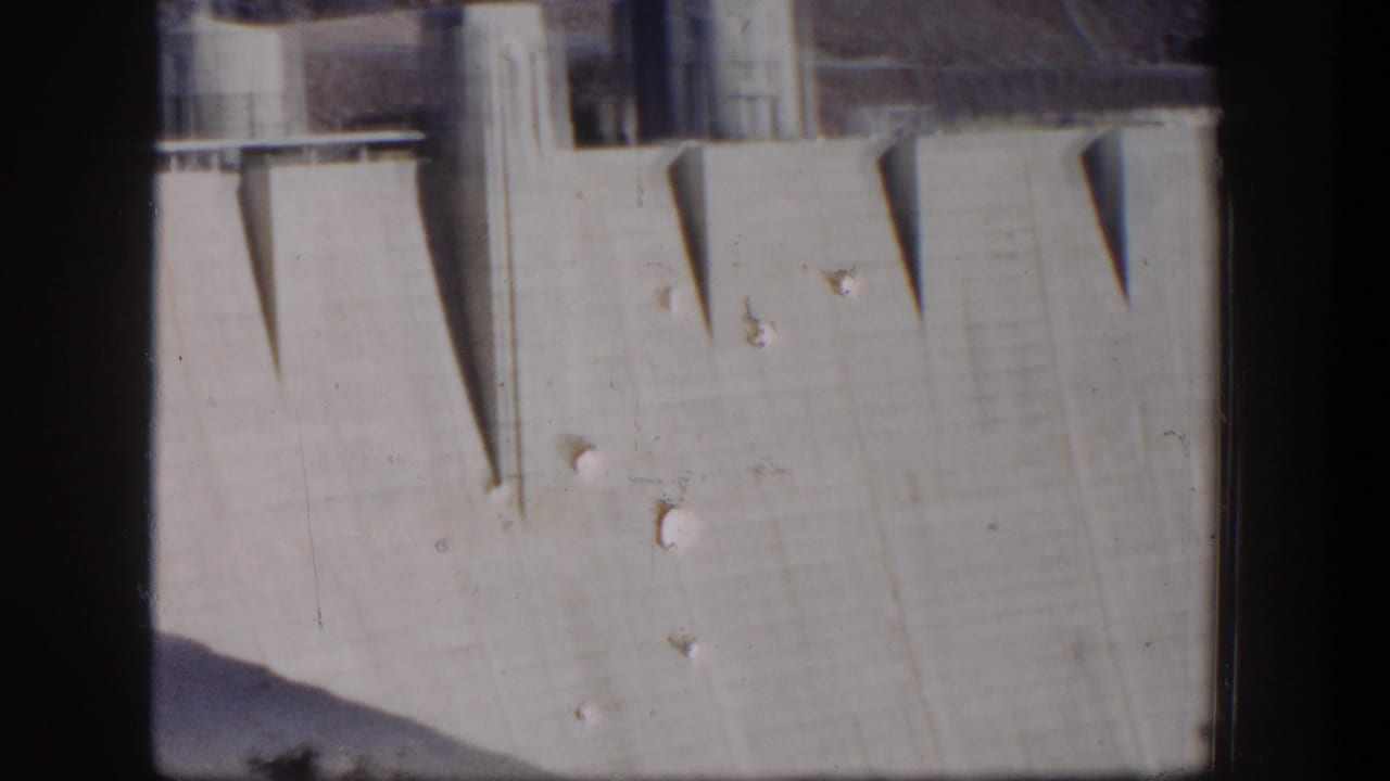1954 Nevada home-movie frame showing a tall concrete dam wall with angular shadows and upper structural details.