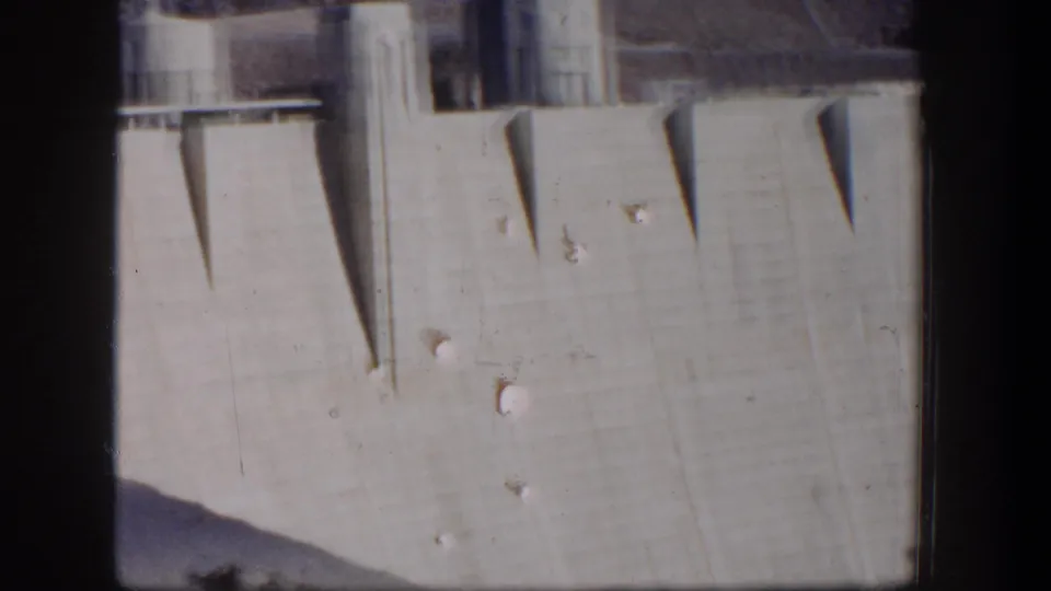 1954 Nevada home-movie frame showing a tall concrete dam wall with angular shadows and upper structural details.