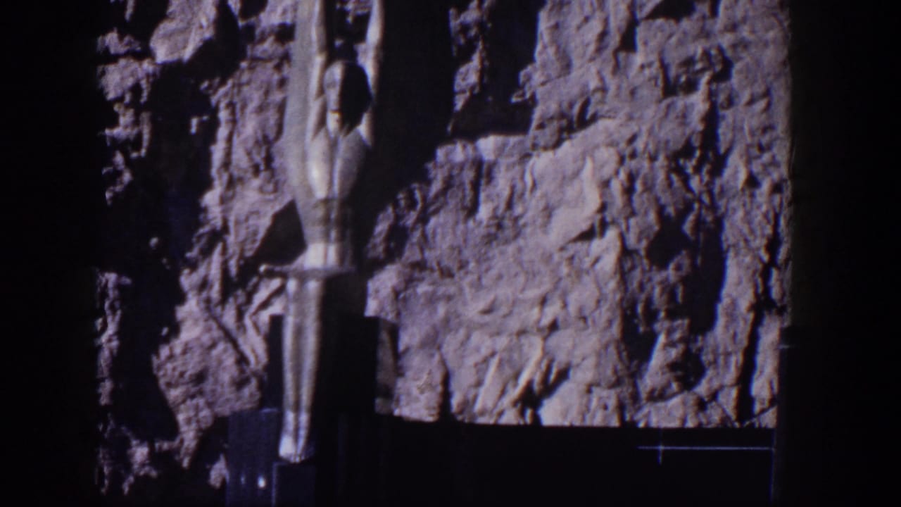 Stylized female sculpture beside a rugged rock face and dark structural base.