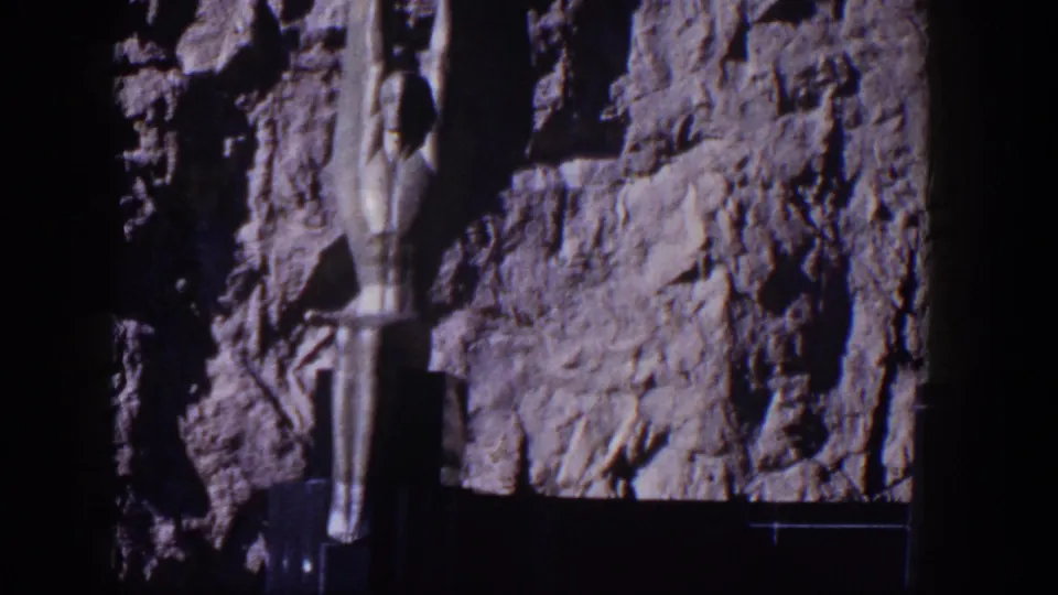 1954 Nevada frame showing a vertical stylized female sculpture set against rough rock near a dam structure.