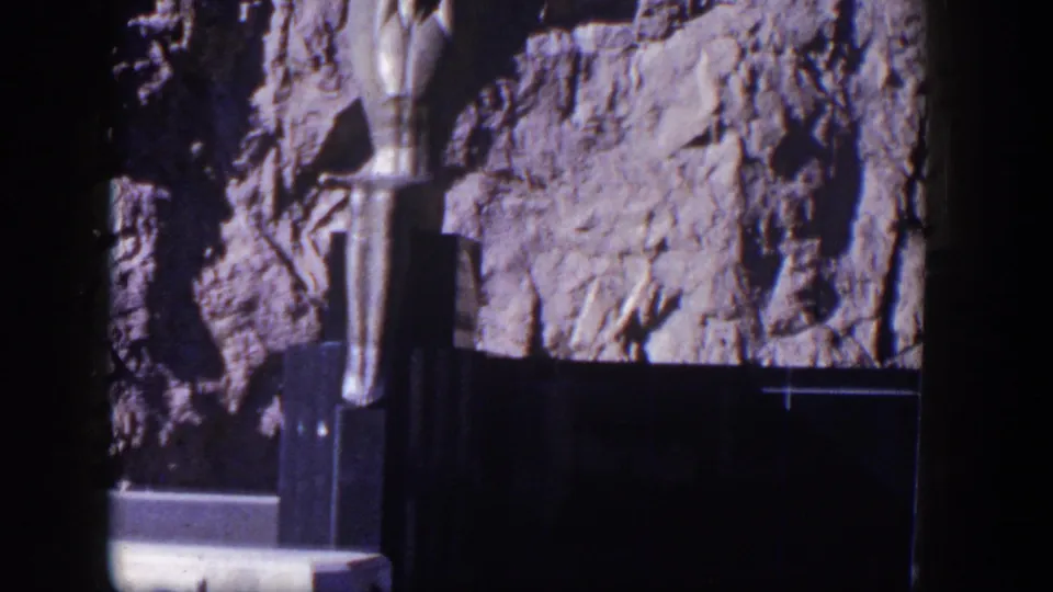 1954 Nevada home-movie frame with a stylized female sculpture, rough cliff surface, and dark base near a dam landmark.