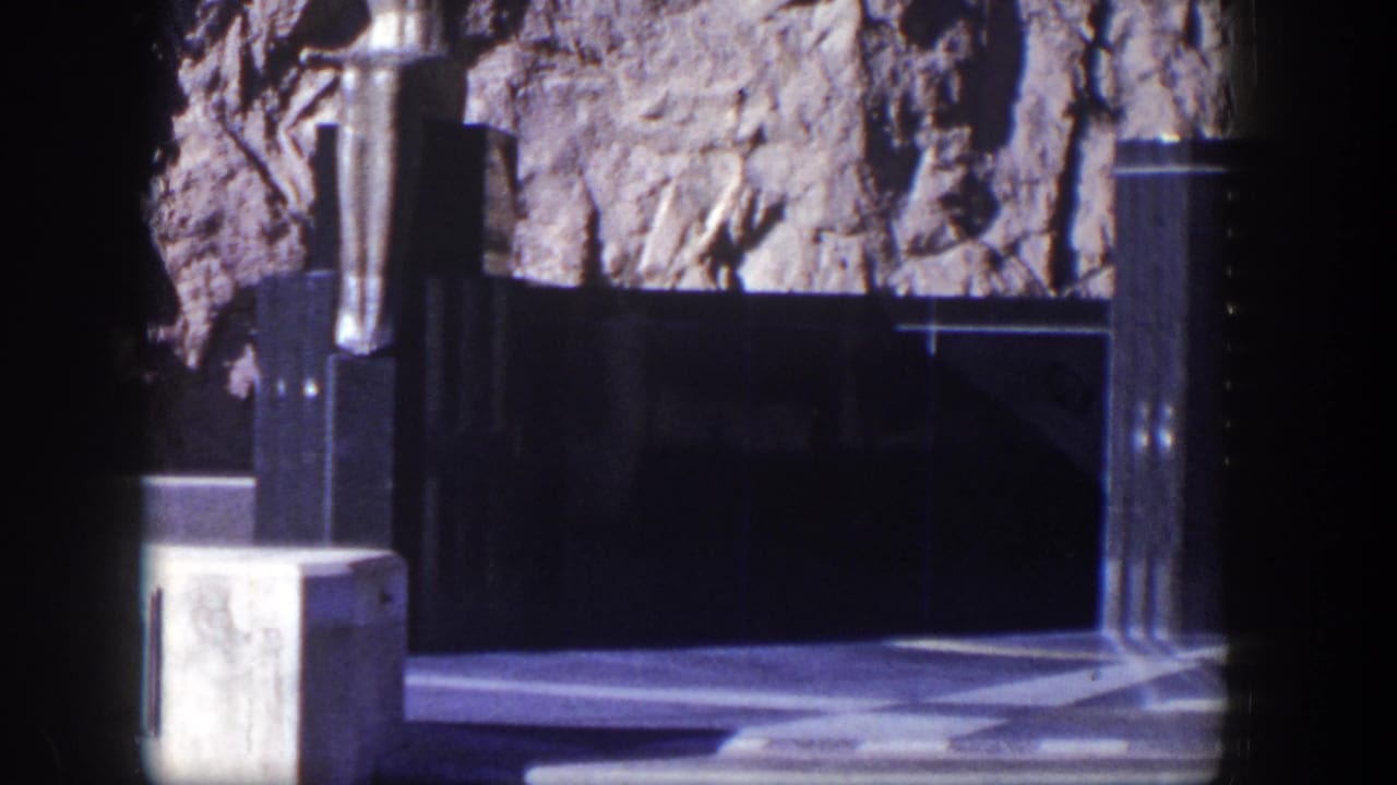 Dark stone memorial structure beside a rugged rock face with checkered paving in bright sun.