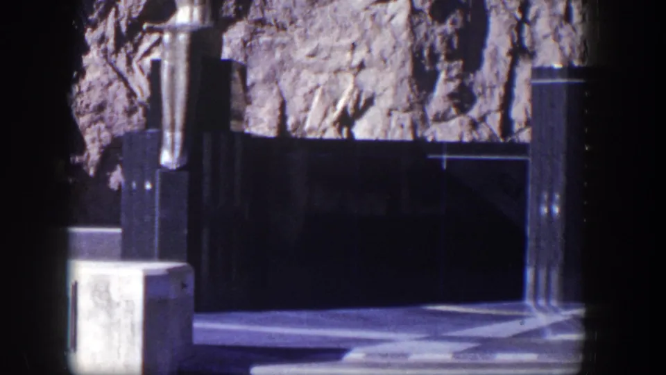 1954 Nevada home-movie frame showing part of a dark stone memorial structure, vertical posts, black panels, and rocky cliff behind.