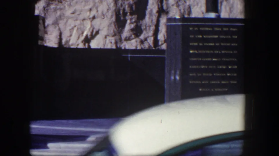 1954 Nevada frame with the engraved memorial partly visible against rock, while a white vehicle passes across the lower right foreground.