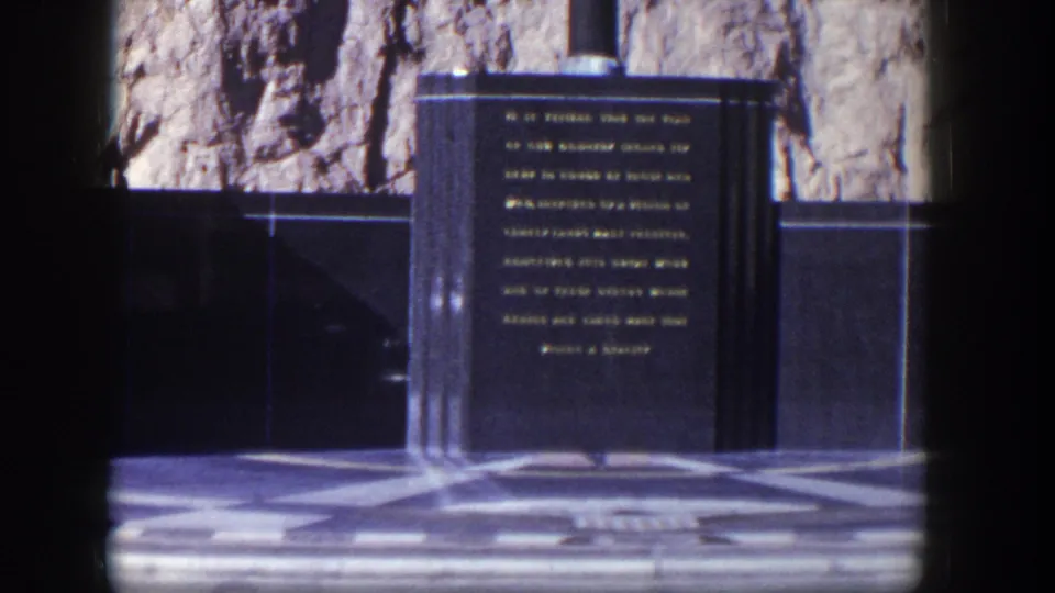 1954 Nevada home-movie frame centered on a dark engraved memorial plaque set before a rugged rock face and patterned paving.