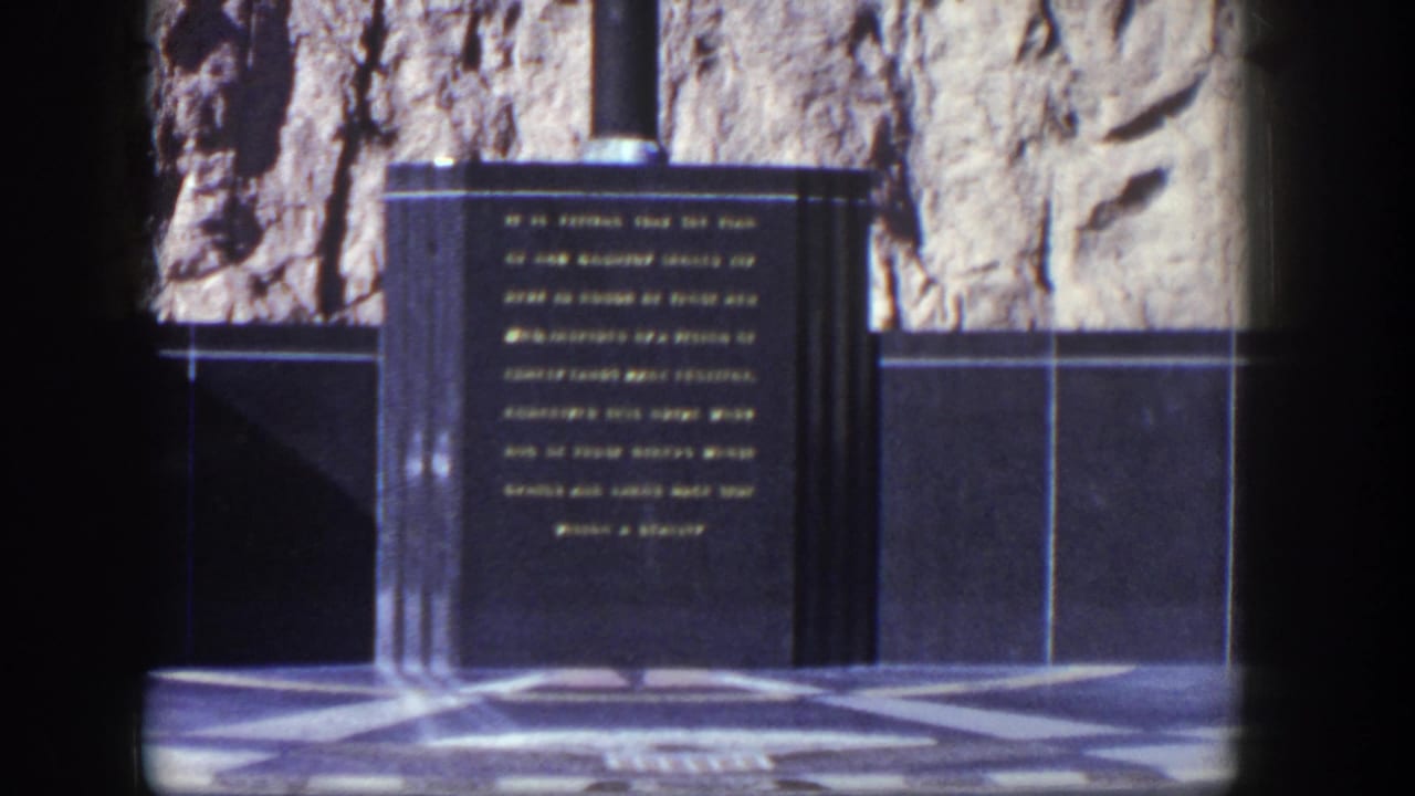 Dark stone monument with gold lettering beside a rough rock wall and patterned paving.
