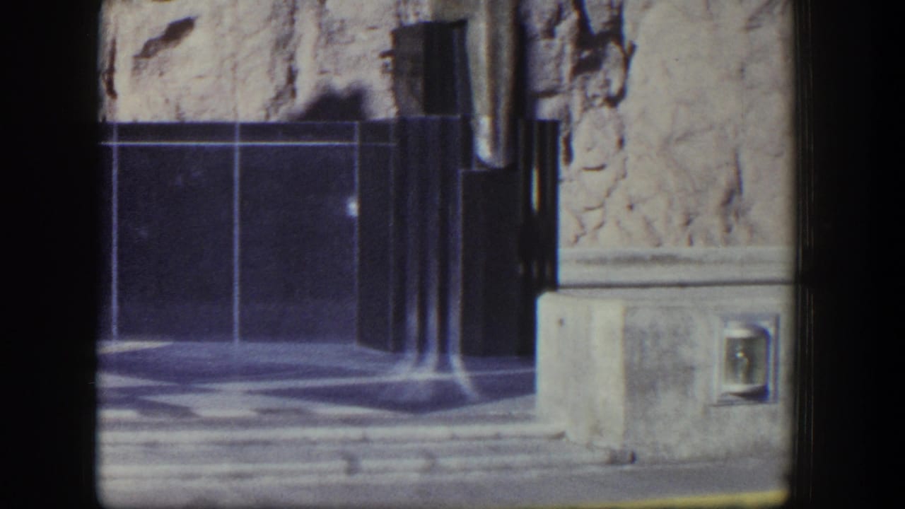 1954 Nevada home-movie frame showing Hoover Dam sculptural stonework, a dark geometric base, rough rock face, and part of a winged figure.