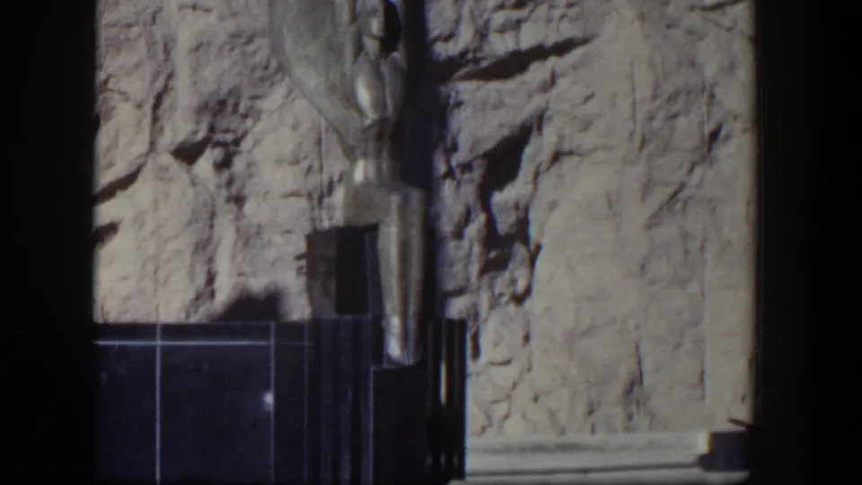 1954 Nevada home-movie view of a Hoover Dam winged figure, with the dark base and textured rock face filling the frame.