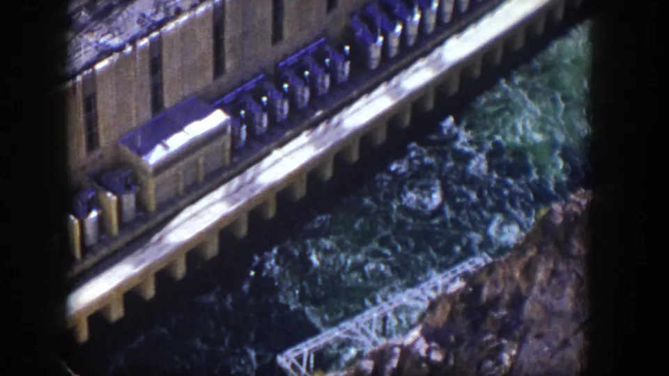 High-angle 1954 Nevada home-movie frame of a dam wall, blue-green spillway water, and a small metal truss below.
