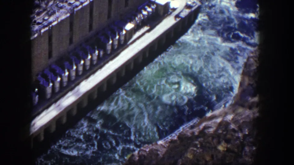 1954 Nevada 8mm still with dam infrastructure above pale blue-green spillway water and rocks.