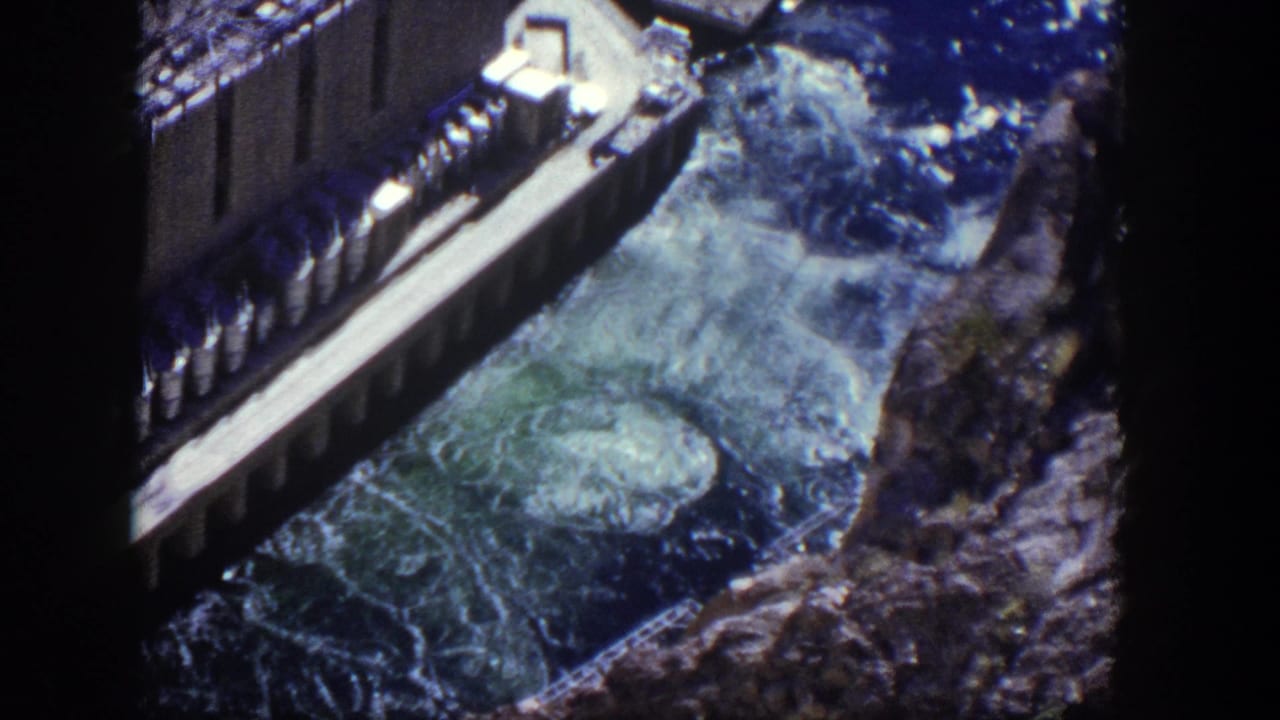 Elevated view of a concrete dam spillway with foaming water between rocky canyon walls.