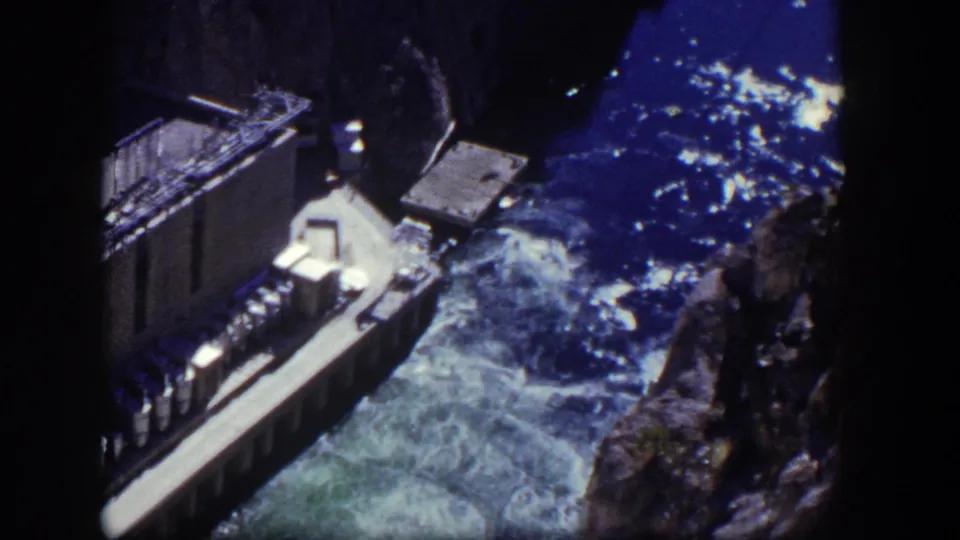 1954 Nevada 8mm frame with a concrete spillway, whitewater turbulence, shadowed dam face, and rugged canyon edge.