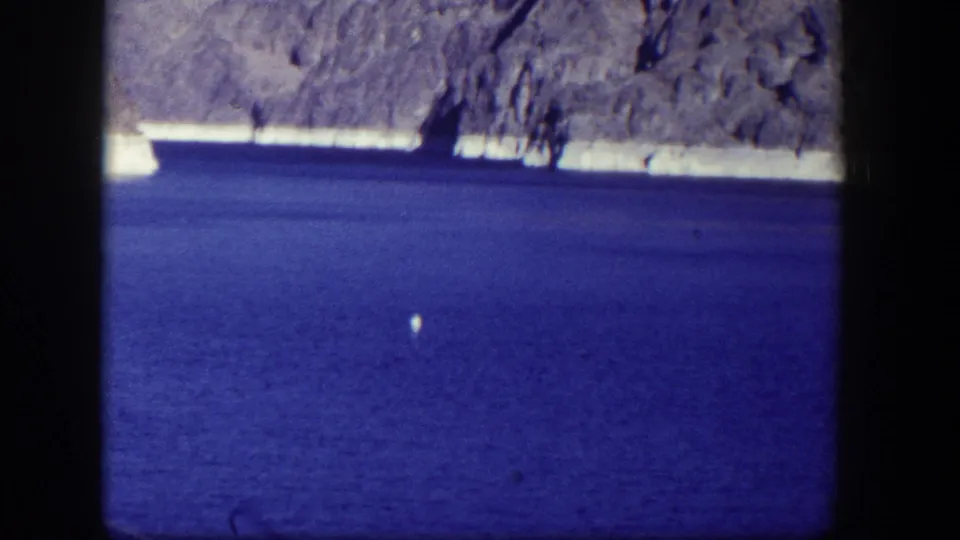 1954 Nevada 8mm view of deep blue reservoir water, pale shoreline, and distant rocky canyon wall, with a small bright speck on the water.