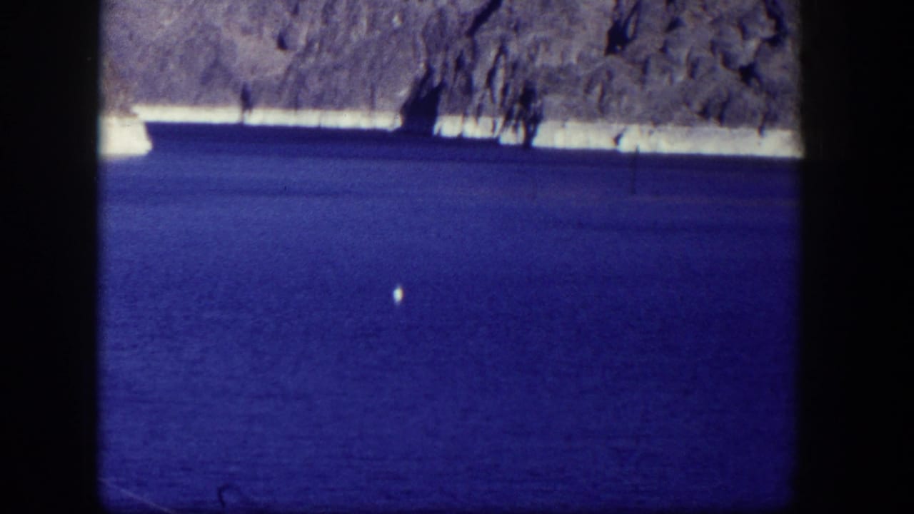 Blue water below a pale rocky shoreline, framed by dark film borders and overhanging branches.