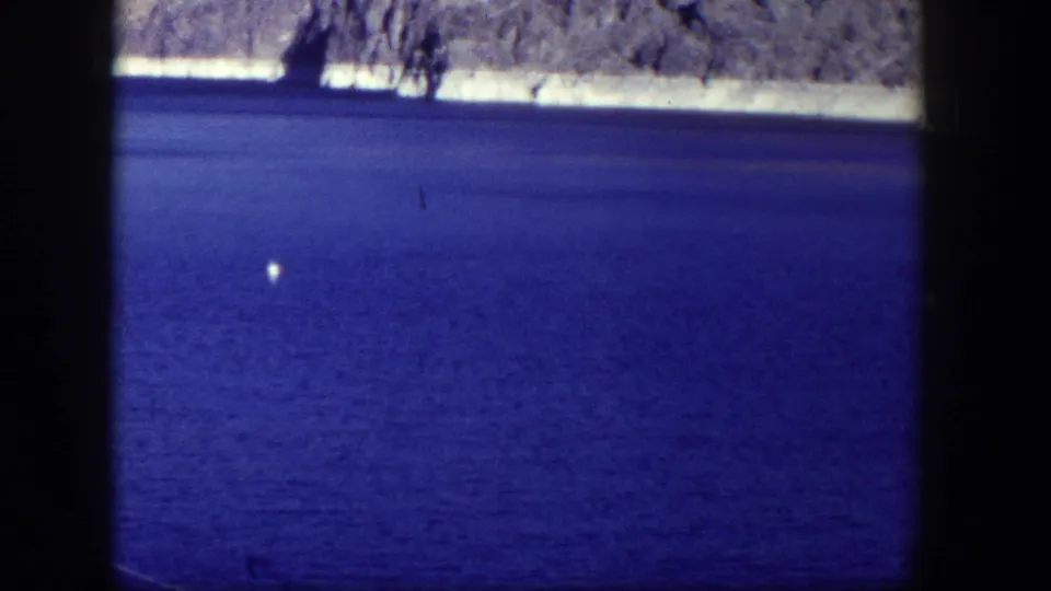 The 8mm frame shows saturated blue water, a distant rocky shoreline, and a small white buoy, with visible film grain and dark borders.