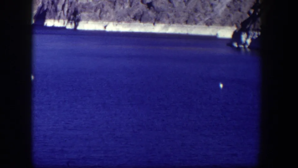 A steady 1954 8mm view of blue water, rocky shore, shaded branches, and a small white buoy near the lower right edge.