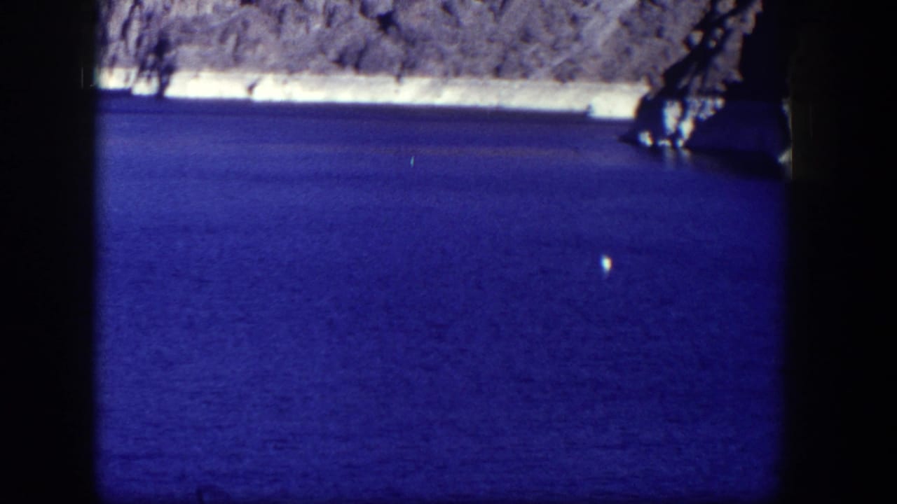 Blue lake water below a rocky shoreline with a dark film border and a small white object floating.