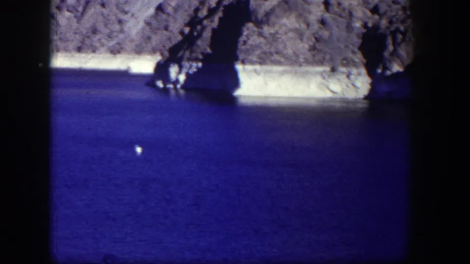 1954 Nevada 8mm view of a rocky, cave-like shoreline over blue water with a small white object on the surface.