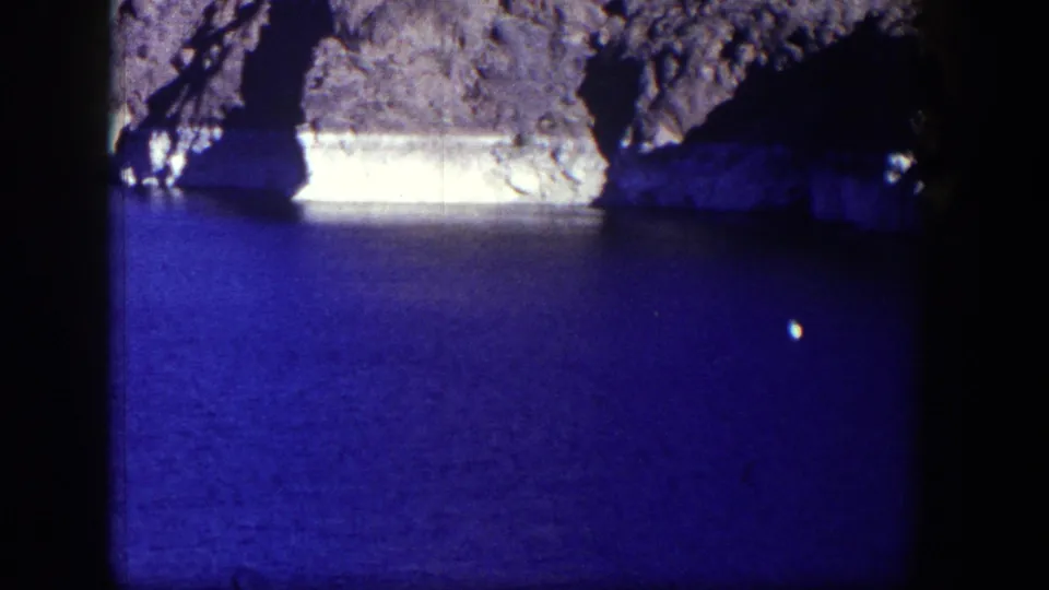 1954 Nevada home-movie frame showing blue lake water, rocky terrain, a dark cave-like recess, and a small white floater.