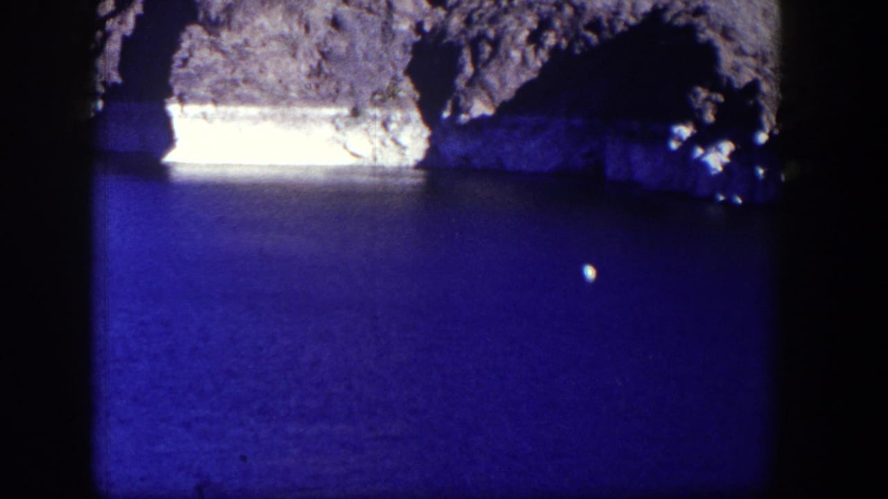 Blue water below a rocky shoreline, framed by dark film edges.