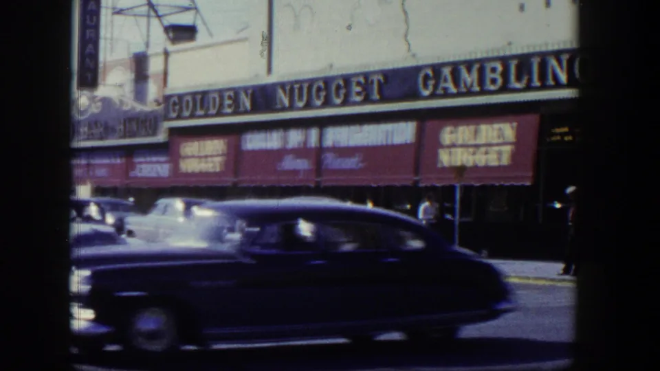 A 1954 Nevada street view shows a dark sedan moving past the Golden Nugget Gambling frontage with awnings and pedestrians nearby.