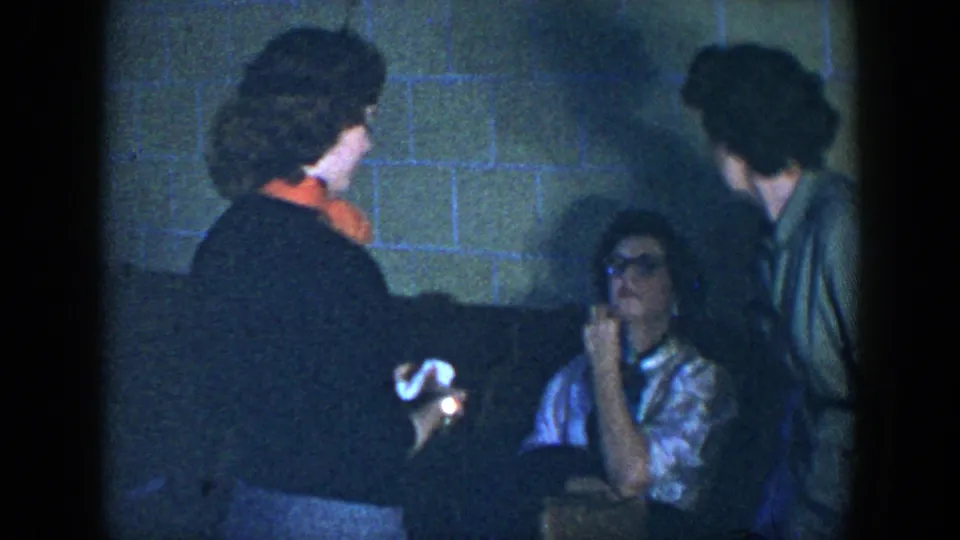 1952 Houston home-movie frame of a woman in dark clothing with an orange scarf holding a handkerchief near seated adults.