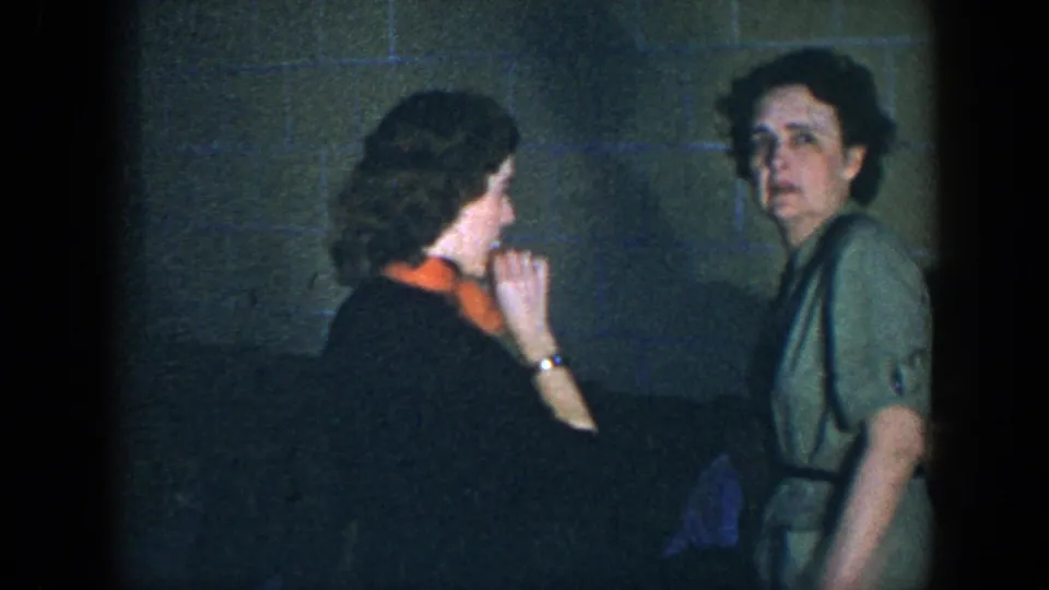 1952 Houston frame of two women near a couch, one in a green dress looking toward the camera and one with an orange scarf in profile.