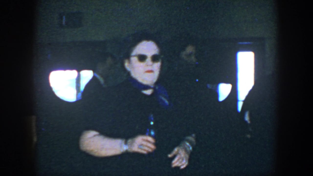 Woman in dark dress and sunglasses holding a bottle in a dim room.