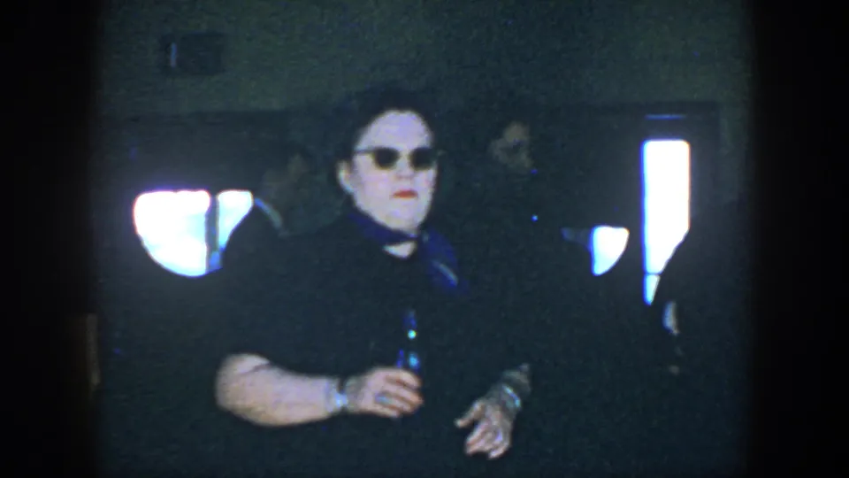 In 1952 Houston home-movie footage, a woman in a dark dress and sunglasses holds a bottle while seated in a dim private interior.