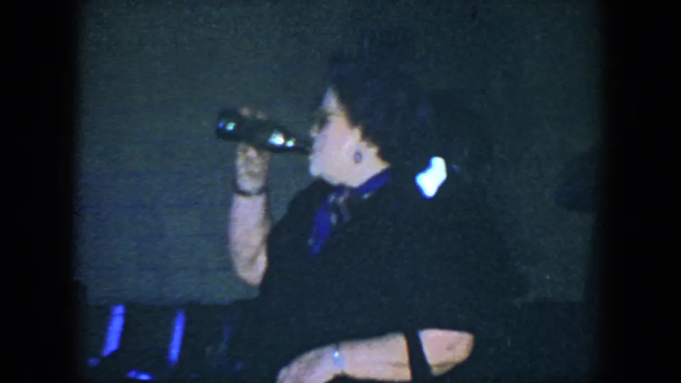 A woman in dark clothing drinks from a bottle in a dim Houston interior, with bright window light behind her.