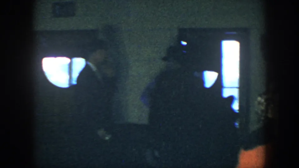 The home-movie frame shows a dim party room with a seated figure, strong window glare, and another person partly entering at the right edge.