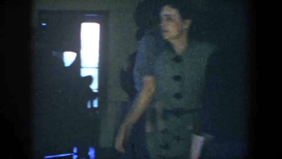 A woman in a patterned dress moves through the dim 1952 Houston party interior near bright doorway or window light.