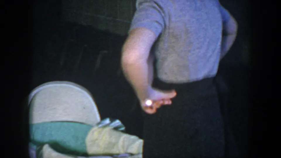 A woman stands near the ruffled baby bassinet in a dim private interior, seen from behind in metadata-supported 1952 Houston footage.