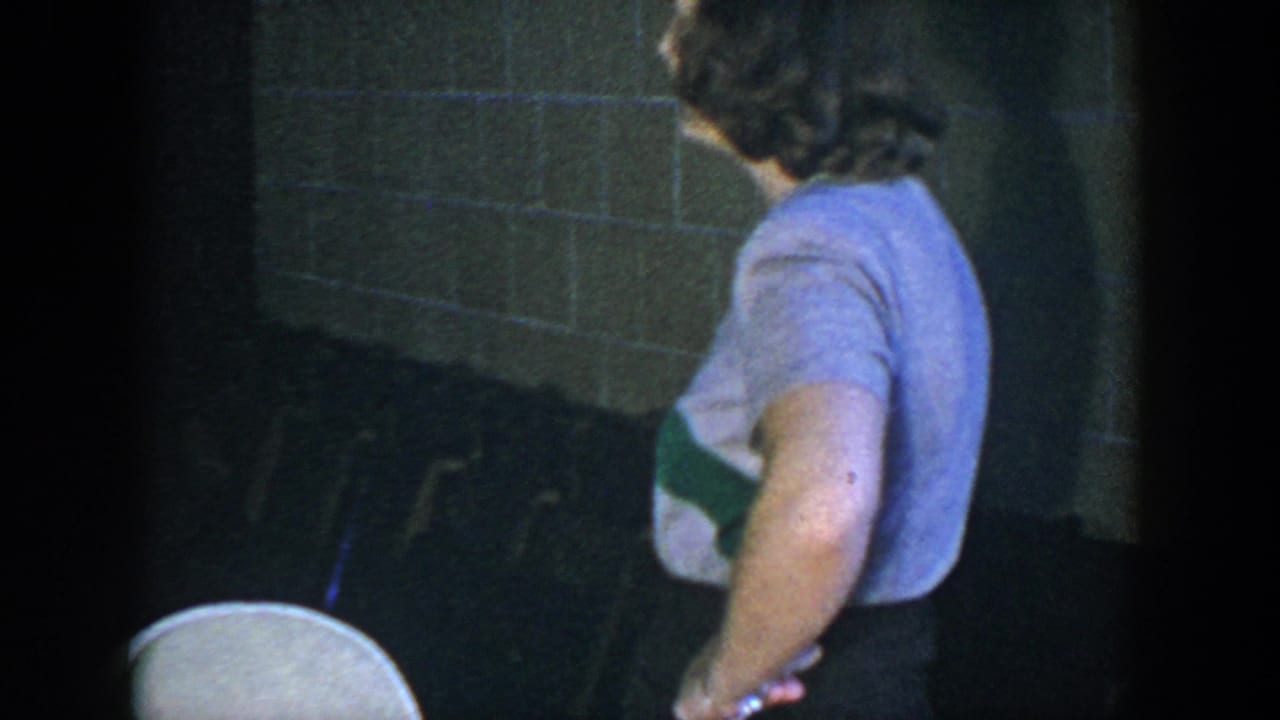 Woman in a light top stands near folding chairs in a dim brick-walled room.
