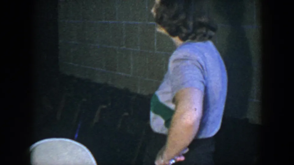 In 1952 Houston home-movie footage, a woman in a light short-sleeved top stands near folding chairs inside a dark brick-walled room.