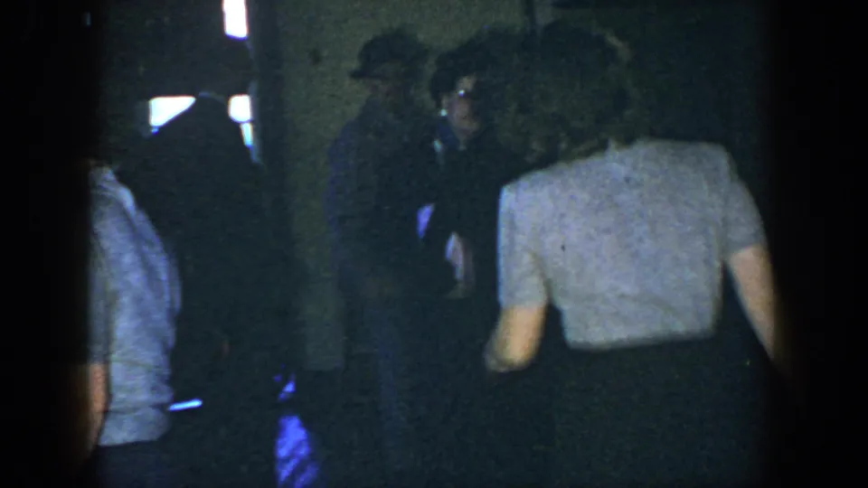 A woman in a light top moves away from the camera near a baby carriage and other adults in the dim Houston home-movie scene.
