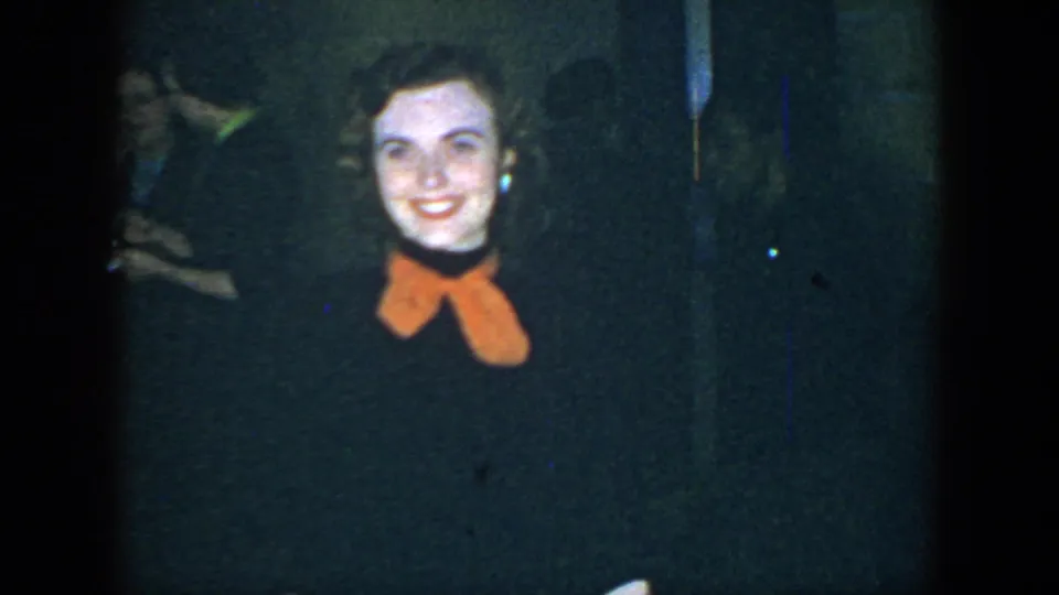 1952 Houston home-movie close view of a smiling young woman wearing a dark dress and bright orange bow in a dim indoor room.