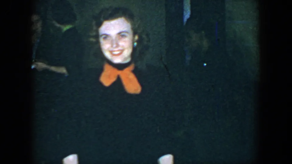 1952 Houston frame of the same smiling young woman in a dark outfit with an orange bow, filmed in soft, grainy 8mm color.
