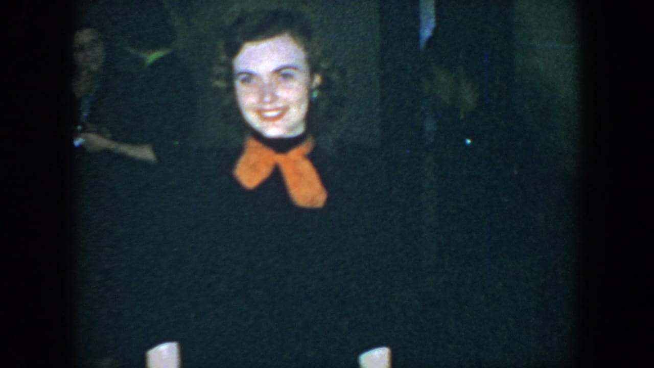 Young woman in dark clothing and orange scarf smiles toward the camera in a dim room.