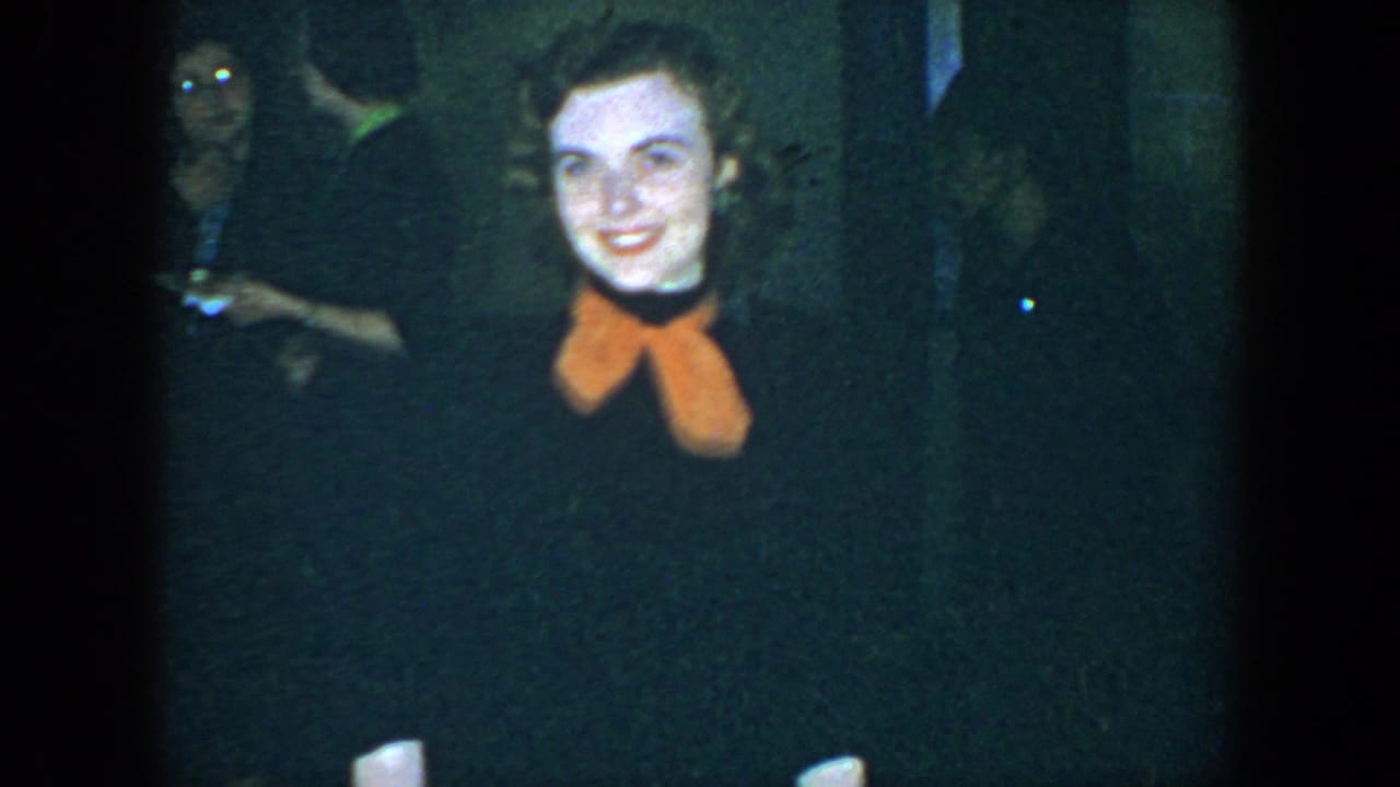 Smiling woman in orange scarf stands before a dark background with another adult behind her.