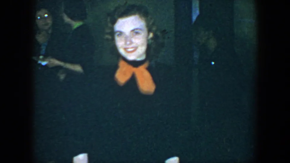 The young woman smiles in a dark indoor Houston scene, with another adult partly visible behind her.