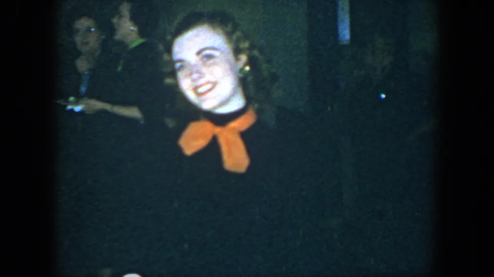 The woman tilts her head and smiles in the final 1952 Houston frame, with indistinct adults in the dark room.