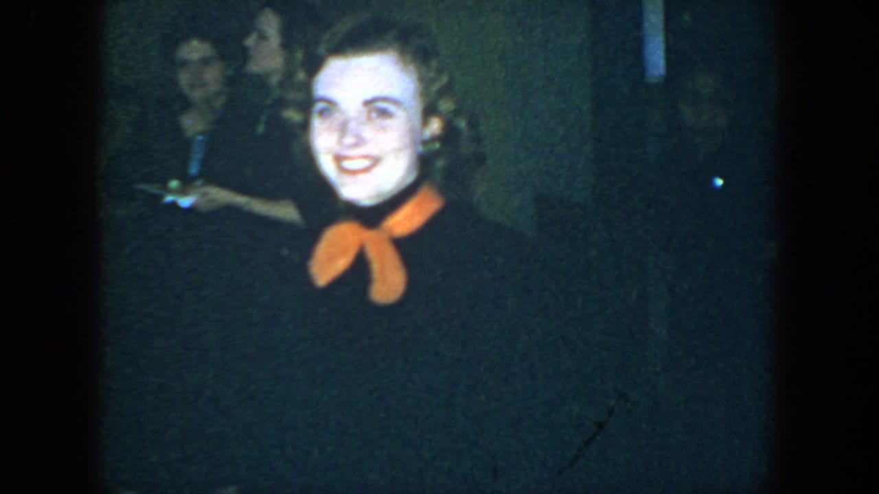 Smiling woman in a dark dress with an orange bow faces the camera in a dim room.