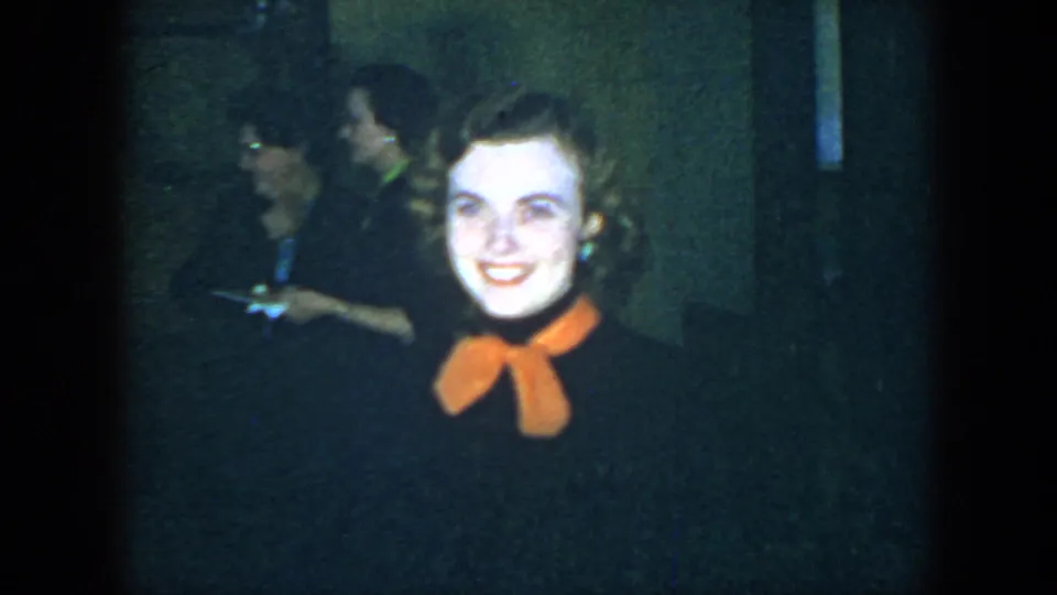 The woman remains centered in the 1952 Houston frame, smiling with an orange neck bow visible and party guests in soft focus behind her.